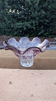 Side view of a purple glass bowl with ruffled edges and clear textured base on an outdoor surface with greenery in the background.