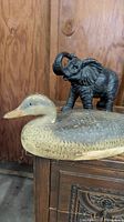 Elephant figurine standing on top of wooden furniture next to a duck decoy with wood panel backdrop.