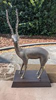 Vintage brass antelope statue standing on a rectangular wooden base. The sculpture showcases the full body in a natural standing pose with long spiral textured horns.