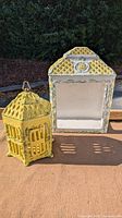 Two yellow vintage ceramic decorative items outdoors: a lantern with lattice design and metal handle, and a wall pocket with floral trims and open front.
