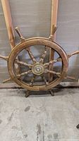 Full view of wooden ship wheel showing eight handles and brass center, placed against a wall on concrete floor.
