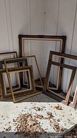 Group view of seven extra-large wooden picture frames of various designs leaning against a wall outdoors with leaf debris on concrete floor.