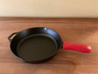 Front-angle view of black cast iron skillet with smooth cooking surface and red handle grip, showing pouring spout and helper handle.