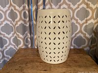 Front view of white ceramic side table with floral pattern cutouts, sitting on wooden surface against curtain backdrop.