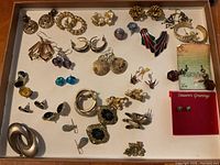 Top-down view of a tray showing a mixture of vintage and modern costume jewelry earrings of various styles and sizes, including studs, hoops, clip-ons, and dangling earrings with a range of colors and motifs.