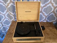 Full view of the Crosley portable turntable with lid open showing the turntable platter, tonearm, and control knobs. The wooden casing and built-in speaker grille at the front are visible.
