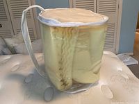 Foam mattress topper rolled and stored inside a transparent plastic carrying bag with white trim and handle, standing upright on a mattress.