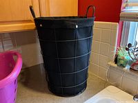 Black fabric laundry basket with metal wire frame, round shape with handles on a countertop next to pink plastic laundry basket.