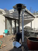Photo of the full standing patio heater on an outdoor wooden deck with a detached metal reflector piece placed on the floor nearby.