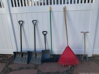 Full view of the lot of outdoor tools including shovels, broom, rakes, and deep watering tool leaning against white vinyl fence with stone and gravel ground.