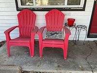 Photo showing two red plastic Adirondack chairs side by side, one with a black and white rabbit patterned pillow, two black metal plant stands, and a red flower pot.