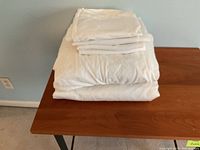 Photo of four white sheets and pillowcases folded stacked on a wooden table next to a light blue wall and a power outlet.