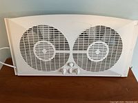 Front view of white plastic dual blade window fan showing two circular fan grilles and three control knobs.