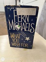 Front cover of 'What You Wish For' by Fern Michaels showing blue dust jacket with white and gold lettering and star designs. Sticker residue and wear visible on top front cover.