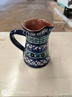 Photo of the ceramic milk jug showing the handle, body and spout, highlighting the dark blue and turquoise painted patterns with a brown glazed interior.