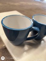 Close-up view of two ceramic coffee mugs with blue exterior and white interior, showing details of the rim and handles.