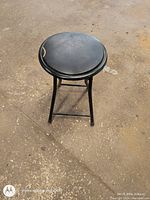 Photo of a black folding stool with a round cushioned seat placed on a textured floor showing the entire stool and its folded-leg design.
