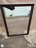 Full view of a rectangular mirror with dark brown wooden frame, standing outdoors against a textured surface.