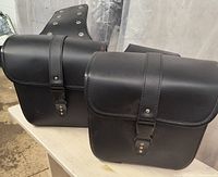 Side view of two black leather motorcycle saddle bags attached to a mounting base with strap closure and rivet details.