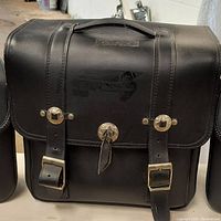 Photo of black leather motorcycle saddle bag showing front with buckles, snap button, and top handle.