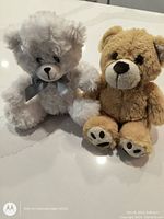Photo of two 8-inch teddy bear stuffed animals, one white with gray ribbon bow and one light brown with white snout and embroidered paw pads, placed on a white surface.