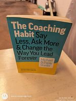 Photo showing the front cover of 'The Coaching Habit' paperback book by Michael Bungay Stanier, held upright on a surface.