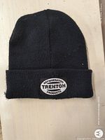 A black knit adult-sized winter beanie with a folded cuff. The cuff features a sewn oval patch with 'Trenton Refrigeration Products' in black and white letters.