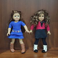 Two American Girl dolls standing side by side in front of wooden background, showing full body and outfit details.