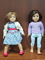 Two American Girl dolls side by side on wooden background. Emily Bennett doll wears blue floral dress, white cardigan, pink shoes. Truly Me doll wears lavender sweater with glitter star, light blue pants, barefoot.