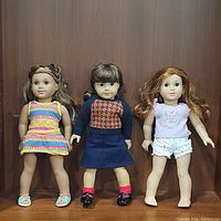 Photo showing three American Girl dolls standing side by side against a wooden background: Molly on left, Lea Clark in the center, and a red-haired doll on the right.