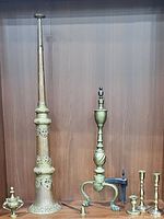 Photo showing all items together: tall Tibetan temple bronze musical instrument with ornate decoration, a bronze brass spiral column andiron with claw feet, three brass candlestick holders, and a brass candle snuffer.