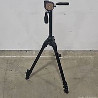 Front view of the SLIK black camera tripod fully opened and standing on the floor showing the three legs and handle.