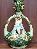 Front view showing the green ceramic vase with two cream-colored handles, decorated with orange-yellow flowers and a hand painted Dutch scene of a man and woman near a windmill.