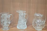 Three glass pitchers arranged on a shelf: cut crystal, etched floral, and clear glass pitchers