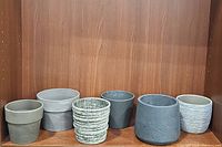 Six assorted grey toned planter pots on wooden shelf, showing variety of textures and shades from light to dark grey.