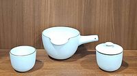 Three white ceramic pieces on wooden background: stick handle batter bowl, lidded sugar dish, and cup with gold rims.