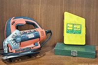 Photo showing Black & Decker orange jigsaw alongside yellow drill set case and green metal socket set box on wooden surface.
