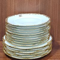 Stack of gold trimmed white porcelain plates showing scalloped and ornately decorated rims, including both dinner and dessert plates stacked separately
