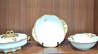 Three Haviland Limoges serving pieces displayed on a wooden shelf: a lidded casserole dish, an oval bowl, and a serving bowl, all white porcelain with gold gilt trim and a monogrammed letter "J".