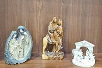 Three nativity statues including a gray-toned resin Holy Family, an olive wood hand-carved Holy Family on a donkey, and a white ceramic nativity votive holder.