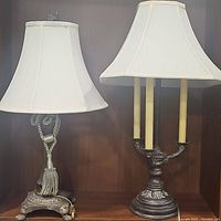 Image showing both the antique style footed lamp and the bronze 3-light lamp side by side, each with white fabric shades, placed on a wooden surface against a dark wood background.