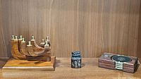 Three items displayed side by side on a wood shelf: a mahogany wood menorah with brass holders, a black stone Chinese chop with an animal figure, and a wooden and brass coaster set.