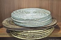 Stack of woven placemats of various colors and textures viewed from the side showing 3 different types stacked: light tan round, dark brown oval, and medium tan round placemats.