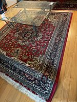 Full angled view of the carpet with a glass table on top, showing its detailed design, rich red background with floral and geometric patterns and fringed ends.
