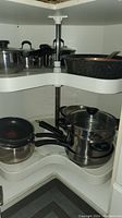 Corner cabinet with two rotating white plastic shelves holding multiple pots and pans including a speckled roasting pan and saucepans with lids.
