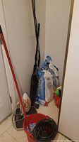 Lot of cleaning supplies including electric iron, mop bucket with wringer and mop, black electric broom, orange pail with cleaning bottles, and packages of PaperTowels brand paper towels in a small corner.