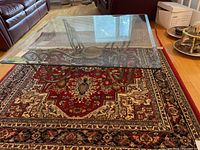 Square glass top coffee table with ornate metal base and gold feet on a red patterned rug.