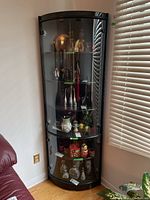 Front view of black curved glass corner cabinet with glass shelves and curved glass front door. Contents inside the cabinet are not included.