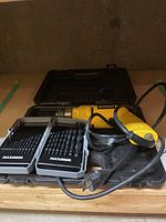 Yellow DeWalt corded drill with power cord placed in open black molded plastic carry case, with two trays of drill bits labeled Maxxomm beside it.