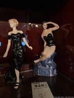 Pair of porcelain figurines inside display case: standing Royal Doulton Alana with blonde hair and black dress to left, seated Chodziez woman in swimsuit on rock to right.
